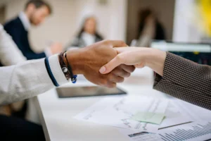 Two people shaking hands over a desk with business documents and a tablet.